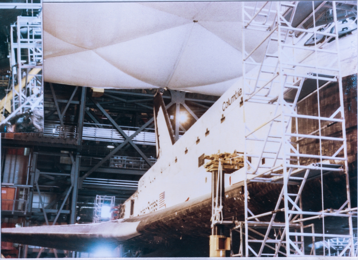 From crew cabin windows in front, to vertical stabilizer in back, the entire right side of Space Shuttle Columbia is visible in this image, taken inside the VAB at Kennedy Space Center, Florida. Note the absence of the right OMS Pod, which was no doubt being serviced elsewhere inside the facility at this time. Above the orbiter, you can see a fabric shroud in place which is there to prevent debris that might drift down from above (at this time, in the mid 1980’s, the VAB was suffering from small bits of concrete that were spalling off from the underside of the roof, and falling onto the interior platforming or sometimes the full 525 feet all the way to the floor in the transfer aisles and high bays), from entering areas where it could become disruptive to critical systems. Behind the orbiter, some of the rigid steel structure of the VAB is visible, along with various items of equipment which it is supporting.